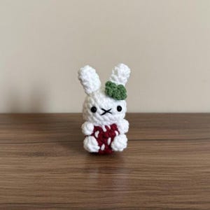 May include: A handmade crochet bunny figurine. The white bunny has a red and white strawberry-like body, black eyes, and a green bow. The amigurumi is displayed on a wooden surface.