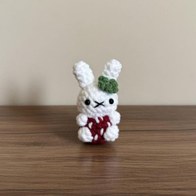 May include: A handmade crochet bunny figurine. The white bunny has a red and white strawberry-like body, black eyes, and a green bow. The amigurumi is displayed on a wooden surface.