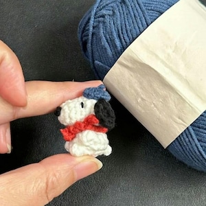 K&ouml;nnte beinhalten: Eine Miniatur-H&auml;kel-Snoopy-Figur, die eine blaue M&uuml;tze und einen roten Schal tr&auml;gt. Der wei&szlig;e K&ouml;rper hat schwarze Ohren und eine schwarze Nase. Im Hintergrund befindet sich ein Kn&auml;uel blaue Wolle.