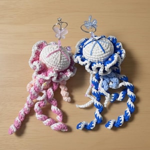May include: Two handmade crochet jellyfish ornaments. One is pink and white, the other is blue and white. Each has a round head, frilly edges, and long, twisted tentacles. They are adorned with small beads and a decorative top.