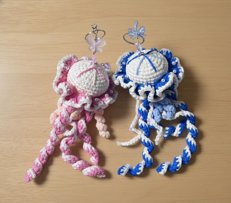 May include: Two handmade crochet jellyfish ornaments. One is pink and white, the other is blue and white. Each has a round head, frilly edges, and long, twisted tentacles. They are adorned with small beads and a decorative top.