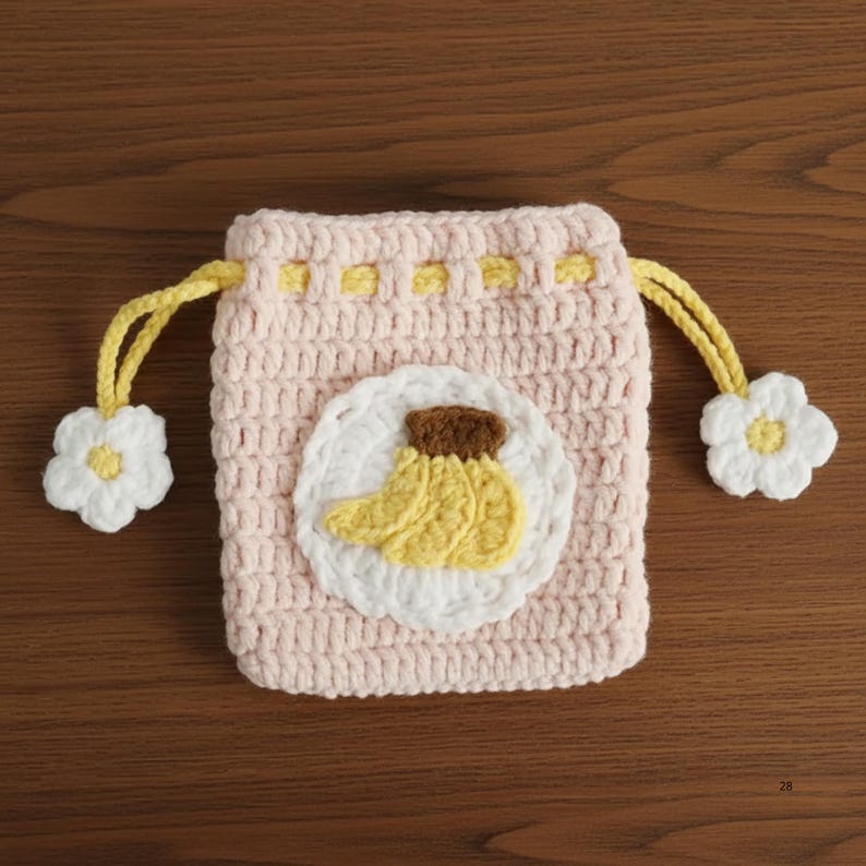 K&ouml;nnte beinhalten: Ein handgefertigter, hellrosa geh&auml;kelter Beutel mit Kordelzug und einem gelb-braunen Bananen-Applikation auf einem wei&szlig;en Kreis. Der Beutel hat gelbe Kordelz&uuml;ge und wei&szlig;e Blumenakzente. F&uuml;r kleine Gegenst&auml;nde.