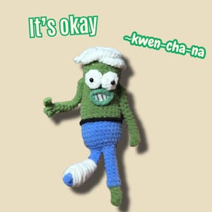 Può includere: Bambola all'uncinetto fatta a mano a forma di mostro verde con cappello bianco, pantaloni blu e gamba fasciata. Il testo "It's okay" e "~kwen-cha-na" sono scritti in verde sopra la bambola.