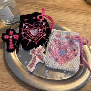 Pode incluir: Bolsas e pingentes de cruz de crochê em preto, branco e rosa, adornados com desenhos de coração e teia de aranha. As bolsas têm cordões e são adornadas com strass. Os pingentes são presos com correntes de prata, apresentados em uma bandeja de prata.