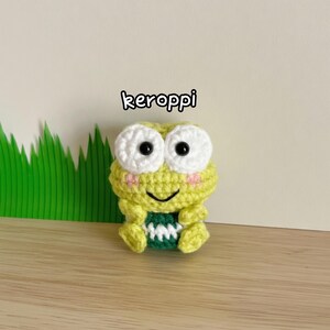 May include: A handmade crochet frog toy in shades of yellow and green, with white eyes and a smiling face. The frog has a green and white patterned belly. The word "keroppi" is written above the frog.