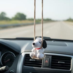 Könnte beinhalten: Ein weiß-schwarzer gehäkelter Hunde-Autoanhänger mit rotem Halsband, der an einem Rückspiegel hängt. Der Hund sitzt auf einer kleinen gehäkelten Schaukel und verleiht dem Innenraum des Autos eine verspielte Note.