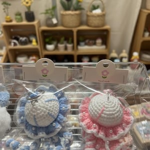 May include: Crocheted jellyfish keychains in blue and pink, with white accents and pearl-like beads. The keychains are packaged in clear plastic, with a small tag that says "hand made". The background shows a display of plants and other crafts.