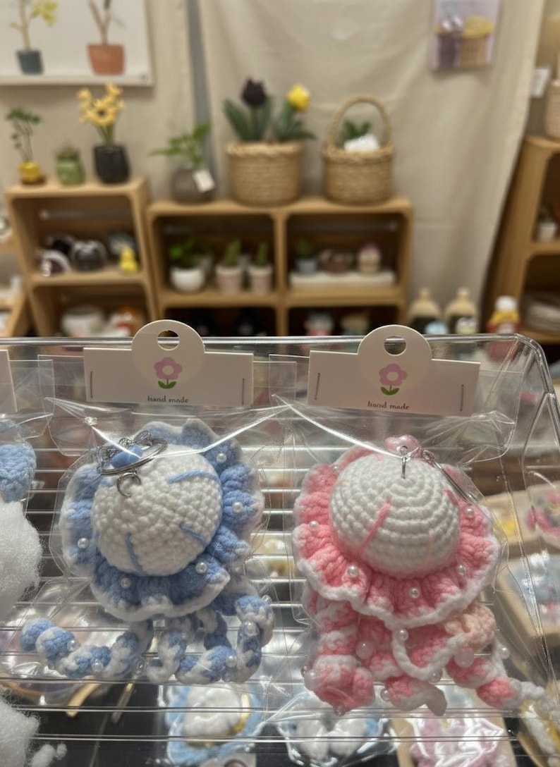 May include: Crocheted jellyfish keychains in blue and pink, with white accents and pearl-like beads. The keychains are packaged in clear plastic, with a small tag that says "hand made". The background shows a display of plants and other crafts.