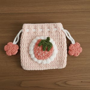K&ouml;nnte beinhalten: Ein hellrosa geh&auml;kelter Kordelzugbeutel mit einem Erdbeer-Applikation. Die Erdbeere hat einen gr&uuml;nen Stiel und wei&szlig;e Samen. Der Beutel hat wei&szlig;e Kordeln und rosa Blumenakzente. Der Beutel ist aus weichem, strukturiertem Garn gefertigt.