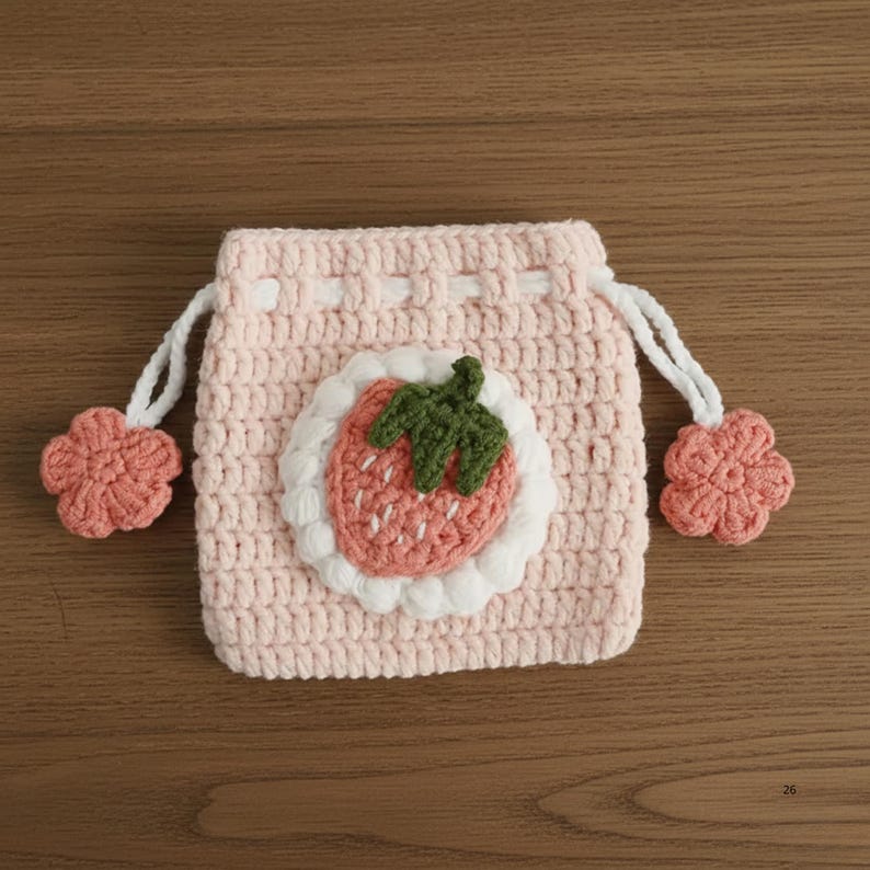 K&ouml;nnte beinhalten: Ein hellrosa geh&auml;kelter Kordelzugbeutel mit einem Erdbeer-Applikation. Die Erdbeere hat einen gr&uuml;nen Stiel und wei&szlig;e Samen. Der Beutel hat wei&szlig;e Kordeln und rosa Blumenakzente. Der Beutel ist aus weichem, strukturiertem Garn gefertigt.