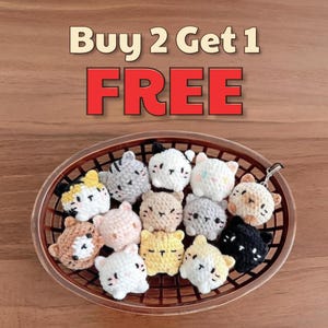 Op de afbeelding: Een verzameling gehaakte katvormige sleutelhangers in een geweven mand. De katten zijn in verschillende kleuren, waaronder wit, grijs, zwart, geel en bruin. De tekst "Buy 2 Get 1 FREE" staat boven de mand.