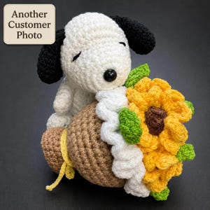 May include: A crocheted white and black dog plush toy holding a brown sack tied with a yellow string, featuring a large yellow sunflower with a brown center and green leaves attached to the side. The text Another Customer Photo is visible.