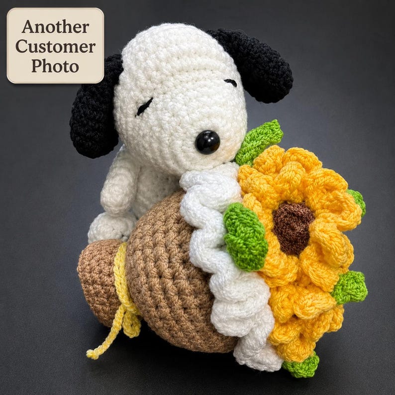 May include: A crocheted white and black dog plush toy holding a brown sack tied with a yellow string, featuring a large yellow sunflower with a brown center and green leaves attached to the side. The text Another Customer Photo is visible.
