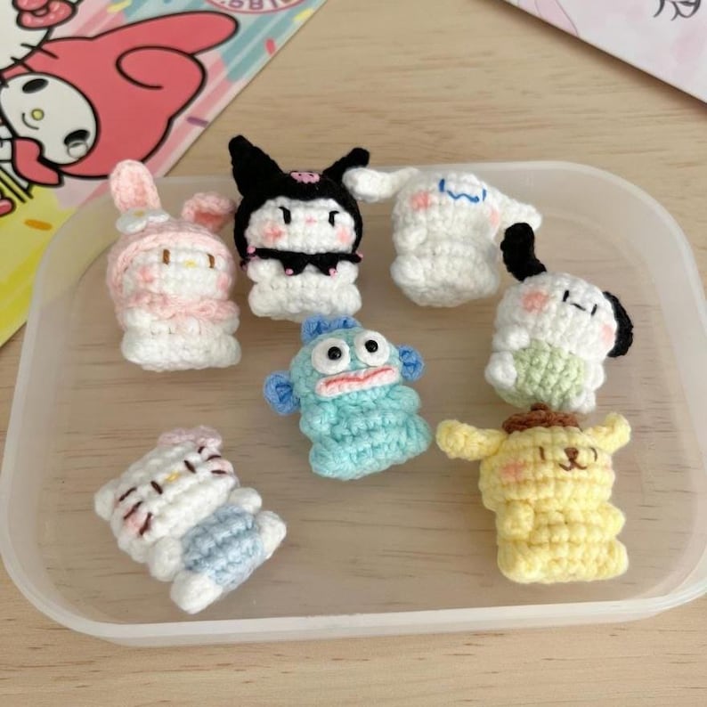 May include: A collection of small, handmade crochet figures in various colors. The figures include a pink bunny, a black and white character, a white ghost, a blue fish, a white and black dog, a yellow dog, and a white cat.