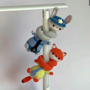 May include: Two crocheted amigurumi toys, a bunny and a fox, clinging to a white pole. The bunny is in blue with a police hat, the fox in orange and yellow. Handmade, colourful, and whimsical.