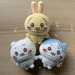Könnte beinhalten: Drei gehäkelte Amigurumi-Puppen: ein gelbes Häschen mit rosa Ohren, ein weißer Bär und eine weiße Katze mit blauen Akzenten. Jede Puppe hat schwarze Augen, rosa Wangen und ein lächelndes Gesicht. Die Puppen sind handgefertigt.