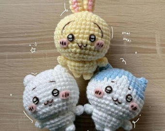 May include: Three crocheted amigurumi dolls: a yellow bunny with pink ears, a white bear, and a white cat with blue accents. Each doll has black eyes, pink cheeks, and a smiling face. The dolls are handmade.