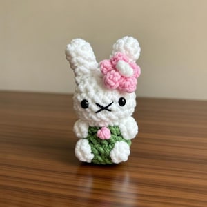 May include: A handmade crochet bunny figurine. The white bunny has black eyes and a stitched 'X' mouth. It wears a green outfit and a pink flower on its head. The bunny is sitting on a wooden surface.