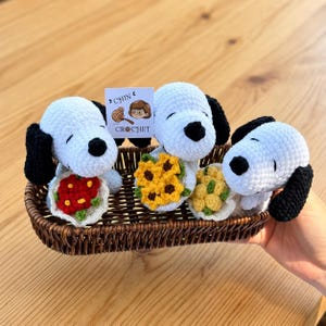 May include: Three white and black crocheted dog figures with floppy ears and black noses, each holding a small crocheted flower bouquet in red, yellow, or light yellow. A small white tag reads CHIN CROCHET with an illustration.