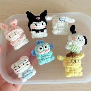 May include: A collection of handmade crochet character figures in a variety of colors. The figures include a pink bunny, a black and white character, a white character with blue accents, a blue character, a white and blue character, a white and black character, and a yellow character.