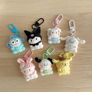 May include: A collection of colorful, crocheted character keychains. The keychains feature various cartoon characters in shades of blue, black, white, pink, and yellow. Each keychain has a metal clasp for easy attachment to keys or bags.