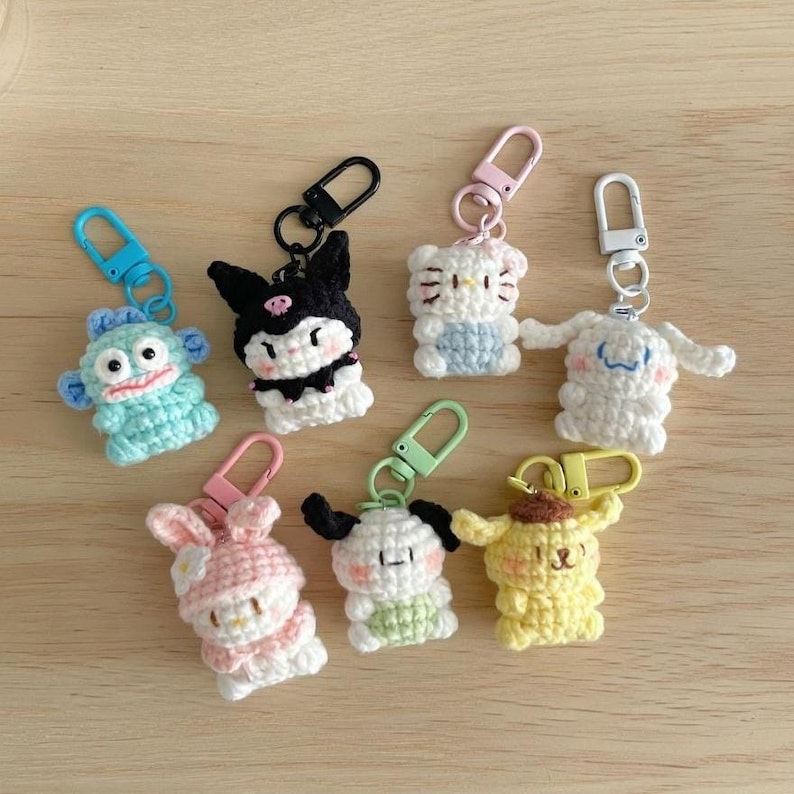 May include: A collection of colorful, crocheted character keychains. The keychains feature various cartoon characters in shades of blue, black, white, pink, and yellow. Each keychain has a metal clasp for easy attachment to keys or bags.