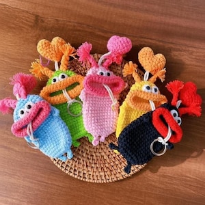 May include: A collection of colorful, crocheted keychains shaped like whimsical creatures. Each keychain features a unique color scheme, including blue, green, pink, yellow, and black, with contrasting pink or orange accents. They have heart-shaped details and key rings.