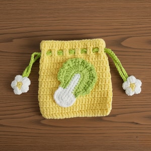 K&ouml;nnte beinhalten: Handgemachte gelbe H&auml;keltasche mit Kordelzug und einem Applikation aus gr&uuml;nem und wei&szlig;em Kohl. Die Tasche hat gr&uuml;ne Kordeln mit wei&szlig;en Blumenakzenten. Die Tasche ist aus strukturiertem Garn gefertigt und liegt auf einer Holzoberfl&auml;che.