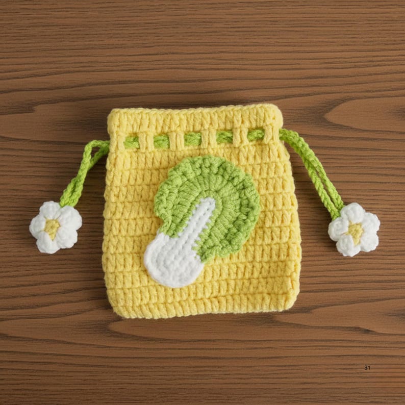 K&ouml;nnte beinhalten: Handgemachte gelbe H&auml;keltasche mit Kordelzug und einem Applikation aus gr&uuml;nem und wei&szlig;em Kohl. Die Tasche hat gr&uuml;ne Kordeln mit wei&szlig;en Blumenakzenten. Die Tasche ist aus strukturiertem Garn gefertigt und liegt auf einer Holzoberfl&auml;che.