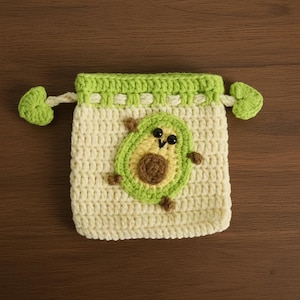 K&ouml;nnte beinhalten: Ein geh&auml;kelter Kordelzugbeutel in Creme mit einem gr&uuml;nen Rand und einem Avocado-Applikation. Die Avocado hat einen braunen Kern, eine gr&uuml;ne Au&szlig;enschicht und kleine schwarze Augen. Der Beutel hat gr&uuml;ne, blattf&ouml;rmige Ziehgriffe.