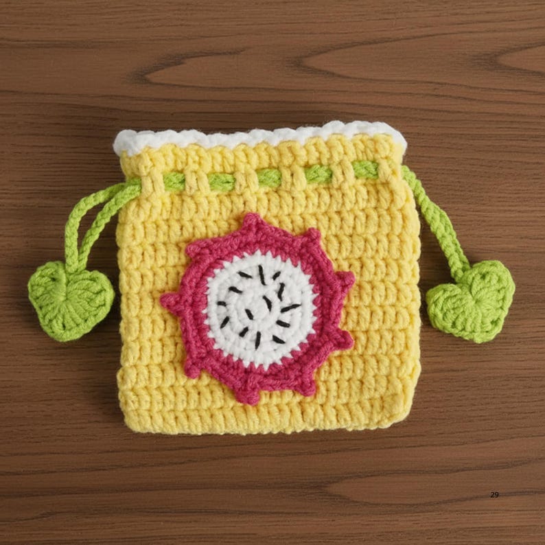 K&ouml;nnte beinhalten: Ein handgefertigter gelber H&auml;kelbeutel mit einem wei&szlig;en und rosa Drachenfrucht-Design. Der Beutel hat einen Kordelzugverschluss mit gr&uuml;nen herzf&ouml;rmigen Akzenten. Der Beutel ist etwa 13 cm breit und 13 cm hoch.