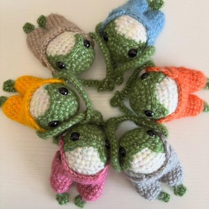 May include: Six handmade crochet frog toys in a circle. Each frog has a green body, white belly, and black eyes. They wear different colored outfits: brown, blue, orange, pink, and gray. The toys are arranged on a white surface.