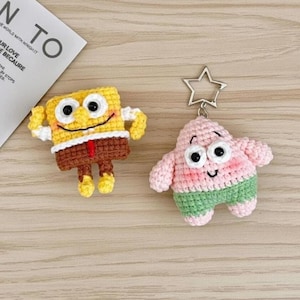 May include: Two handmade crochet keyrings featuring cartoon characters. One is yellow and brown, resembling a sponge, and the other is pink and green, resembling a starfish. Both have black eyes and smiling faces. A silver star-shaped keyring is attached to the starfish.