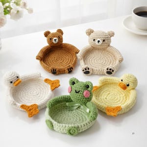Può includere: Una collezione di cinque vassoi fatti a mano all'uncinetto a forma di animali. Il set include un orso marrone, un orso beige, un'anatra bianca, una rana verde e un'anatra gialla. Ogni vassoio ha una base circolare.