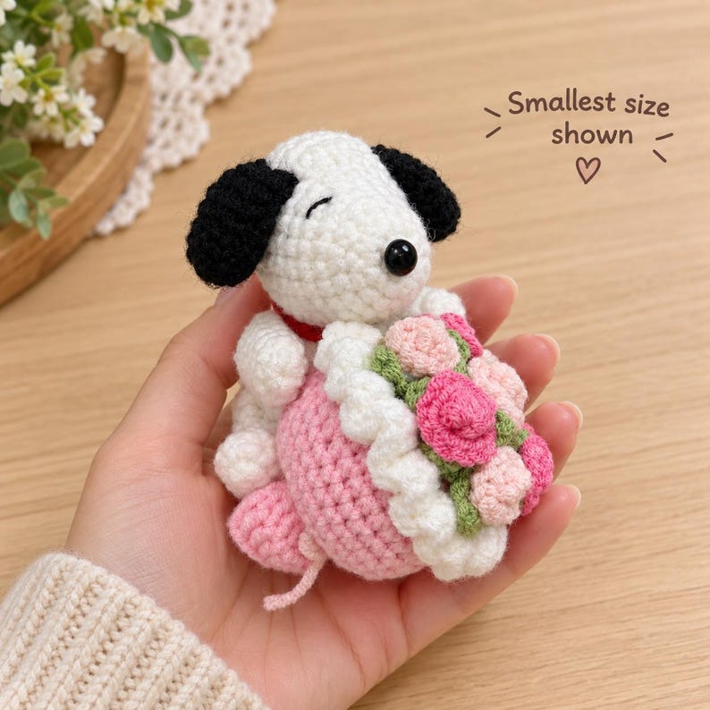 May include: A white and black crocheted dog figurine with a red collar, holding a bouquet of pink and light pink roses with green leaves. The text "Smallest size shown" with a heart symbol is printed in the upper right corner.