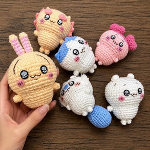 May include: A collection of six small, handmade crochet plush toys featuring various pastel colors including peach, white, pink, and blue. Each plush has embroidered facial features with large black eyes, pink blush marks, and small limbs.