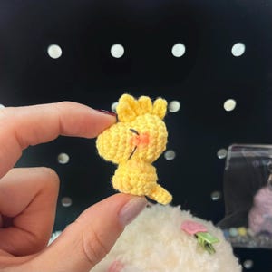 K&ouml;nnte beinhalten: Ein kleiner, gelber, geh&auml;kelter Vogel mit einem l&auml;chelnden Gesicht und orangefarbenen Wangen. Der Vogel hat einen kleinen Schwanz und eine Federkrone auf dem Kopf. Der Vogel wird zwischen zwei Fingern gehalten.