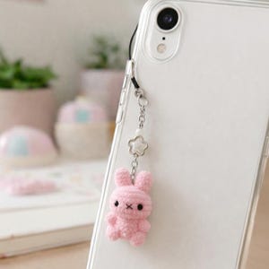May include: A pink crochet bunny phone charm attached to a clear phone case. The bunny has black eyes and a small X for a mouth. The charm is connected to the phone case with a silver chain and a star-shaped charm.