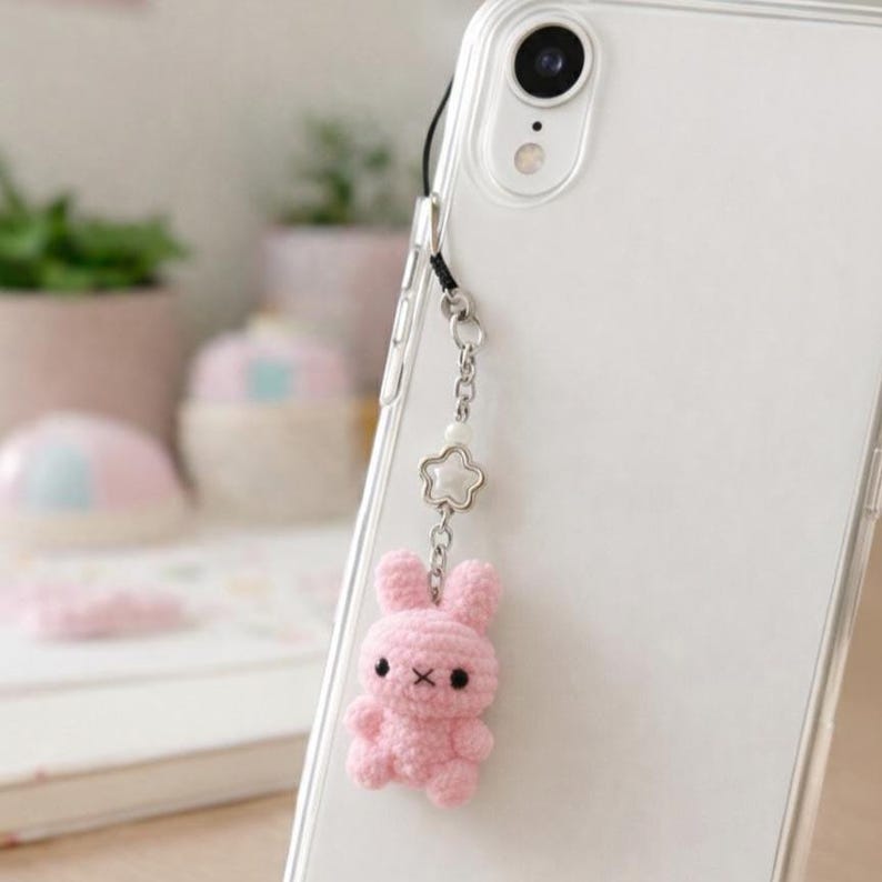 May include: A pink crochet bunny phone charm attached to a clear phone case. The bunny has black eyes and a small X for a mouth. The charm is connected to the phone case with a silver chain and a star-shaped charm.