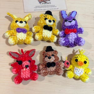 Puede incluir: Seis peluches de ganchillo hechos a mano en amarillo, morado, rojo y marrón. Los juguetes incluyen un conejo, un oso, un zorro y un pollito, cada uno con características únicas como pajaritas y sombreros. Dispuestos sobre una superficie de madera clara.