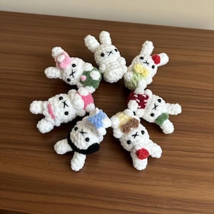 May include: A collection of seven white crochet bunny figures, each with unique decorative accents. Some bunnies feature bows, while others have colorful details like a red heart or a blue hat. The bunnies are arranged in a circular pattern.