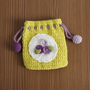 K&ouml;nnte beinhalten: Ein handgefertigter gelber H&auml;kelbeutel mit Kordelzug und einer wei&szlig;en Blumenapplikation. Die Blume hat lila und gr&uuml;ne Akzente. Die Tasche hat lila Kordelz&uuml;ge mit passenden Pompons. Die Tasche liegt auf einer Holzoberfl&auml;che.