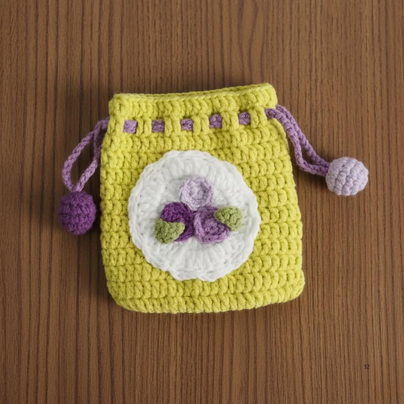 K&ouml;nnte beinhalten: Ein handgefertigter gelber H&auml;kelbeutel mit Kordelzug und einer wei&szlig;en Blumenapplikation. Die Blume hat lila und gr&uuml;ne Akzente. Die Tasche hat lila Kordelz&uuml;ge mit passenden Pompons. Die Tasche liegt auf einer Holzoberfl&auml;che.