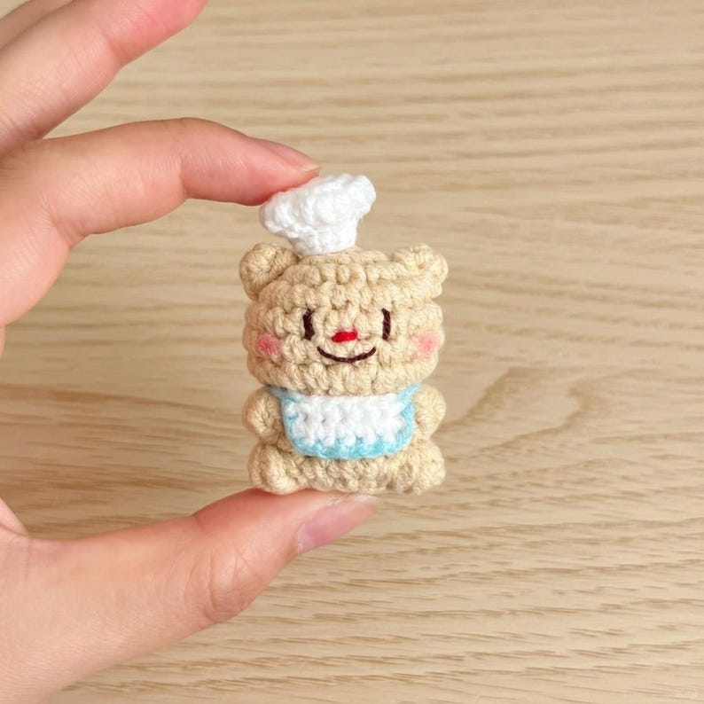 May include: A crocheted bear figurine wearing a chef's hat and a bib. The bear is tan with pink cheeks, a red nose, and a smiling mouth. The bib is white with a light blue trim. The chef's hat is white.