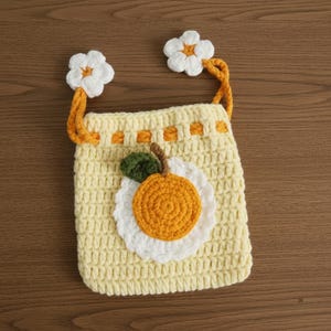 K&ouml;nnte beinhalten: Handgefertigte H&auml;keltasche in Hellgelb mit einem gro&szlig;en Orangen-Applikation mit wei&szlig;em, gewelltem Rand und gr&uuml;nem Blatt. Die Tasche hat zwei wei&szlig;e Blumenakzente und orangefarbene Tr&auml;ger.
