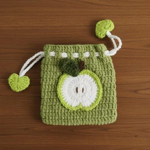 K&ouml;nnte beinhalten: Ein handgefertigter gr&uuml;ner Kordelzugbeutel aus H&auml;kelarbeit mit einem wei&szlig;en Apfelscheiben-Design. Der Apfel hat eine gr&uuml;ne Umrandung, eine wei&szlig;e Mitte und einen braunen Stiel. Die Tasche hat einen wei&szlig;en Kordelzug und zwei gr&uuml;ne Blattakzente. Die Tasche liegt auf einer Holzoberfl&auml;che.
