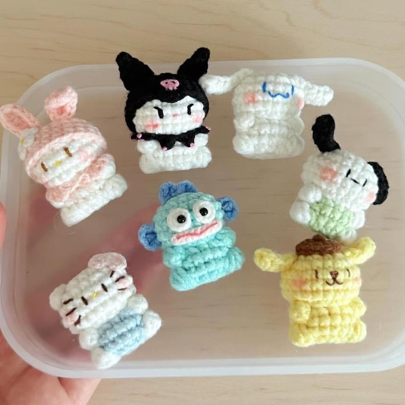 May include: A collection of seven crocheted character figures in a clear plastic container. The figures are in various colors, including pink, white, black, blue, and yellow. The characters appear to be from a popular cartoon series.