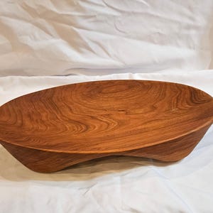 Puede incluir: Un cuenco de madera hecho a mano con una forma orgánica única. El cuenco está hecho de madera de color marrón rojizo cálido, que muestra los patrones naturales de la veta. El diseño del cuenco es funcional y decorativo, adecuado para diversos usos.