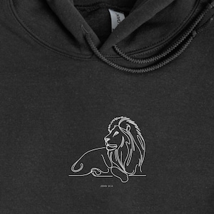 May include: A black hoodie with a white line drawing of a lion. The lion is lying down with its head turned to the side. The hoodie has a drawstring hood and long sleeves. The image is on a white background.