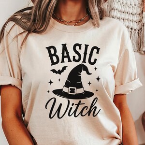 May include: A beige short-sleeve t-shirt featuring a black graphic print. The design includes the text "BASIC Witch" in a stylized font, accompanied by a central witch hat, a small bat silhouette, and several four-pointed star accents.